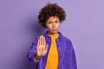 Serious annoyed dark skinned Afro American woman keeps palm in stop gesture asks not to bother her looks angrily wears purple jacket expresses restriction or denial. Do not come closer please - obrazy, fototapety, plakaty