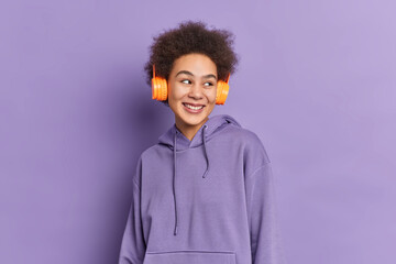 Horizontal shot of curly pretty teenage girl wears stereo headphones listens audio track looks happily aside dressed in sweatshirt isolated over purple background. Monochrome shot. Lifestyle