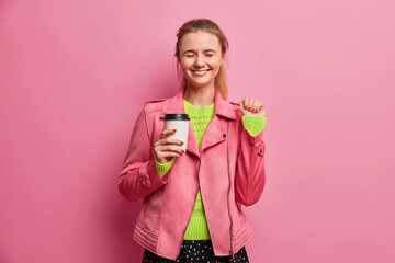 Happy joyful millennial girl raises hand drinks coffee to go closes eyes from happiness wears stylish jacket enjoys free time isolated on pink background. Overjoyed female model with aromatic beverage