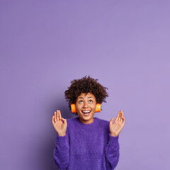 Vertical shot of joyful curly haired pretty woman raises palms and looks with interest at ceiling listens music via wireless headphones dressed in casual sweater isolated over purple background.