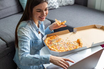 A young smiling woman is working on her laptop and eating pizza at home.