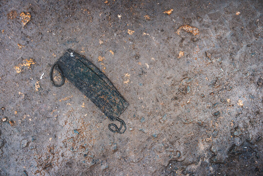 Used trampled dirty fabric face mask lying on muddy ground. Public spaces are flooded with discarded protective equipment.