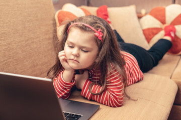 Little girl lying on sofa uses laptop for distance learning or for entertainment