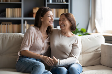 Overjoyed young woman with mature mother chatting, laughing at joke, enjoying pleasant conversation, middle aged mum and grownup daughter hugging, having fun, sitting on cozy couch at home - obrazy, fototapety, plakaty