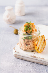 Delicious homemade chicken pate with spices and dill in a glass jar and a slice of bread nearby and caper on a light board background. Selective focus.