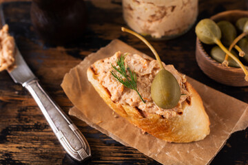 Sandwich with delicious homemade chicken pate with spices and dill in a glass jar and a slice of bread nearby and caper served in vintage style on old board and background.Selective focus.