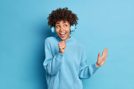 Happy ethnic female sings favorite song keeps hand near mouth as if microphone wears wireless headphones keeps palm raised dressed in knitted sweater isolated over blue background listens music - Powered by Adobe