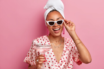 Positive African American woman laughs happily wears sunglasses bath towel dressing gown holds glass of martini cocktail has fun on domestic party smiles broadly isolated over pink background - obrazy, fototapety, plakaty