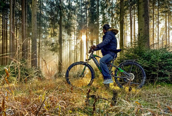 Naklejka premium Mountain biker cyclist man is looking in the forest for the sunbeams coming through the trees, idyllic moment in the morning.