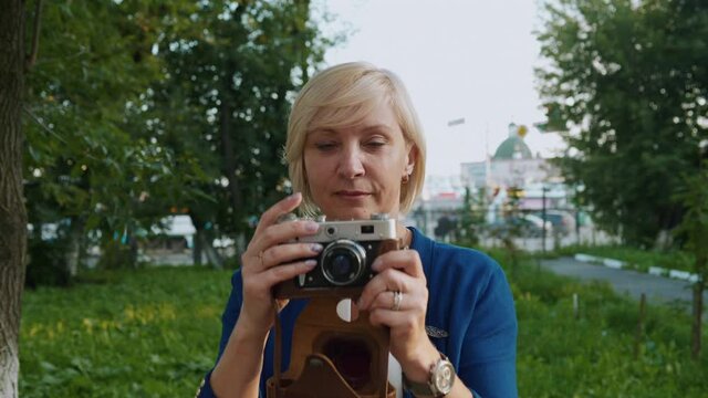 A middle-aged blonde taking photos by vintage film photo camera in park on urban background.