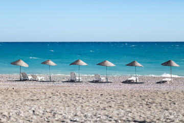 Beautiful view of beach with pebbles and blue ocean or azure sea with white sun loungers and wicker umbrellas. Concept of postcard about vacation, rest in paradise, travel.
