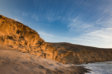 Brightly lit by the rising sun, a cliff of yellow sandstone on the seashore with large cracks and smooth lines of erosion. Thin threads of cirrus clouds in the blue sky. No people