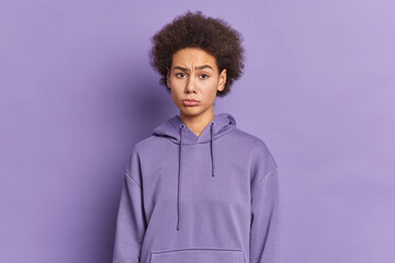 Fototapeta na wymiar Beautiful curly sad teenage girl looks unhappily at camera dressed in hoodie being offended at someone isolated over purple background hears bad words has problems. Negative emotions concept