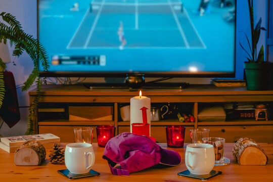 Cozy living room scene with burning candles, coffee mugs and decorative winter objects with a hard court tennis match on TV in the background. Selective focus on candles, TV image is blurred.