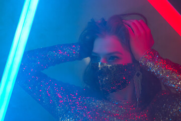 Woman wearing stylish sequin face mask posing, dancing in colorful bright neon uv blue and red lights. Fashion during quarantine of coronavirus outbreak