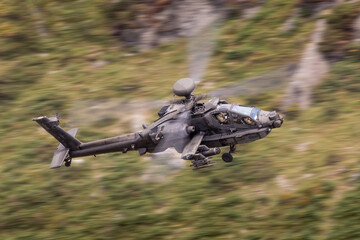 Low Flying AH-64 Apache attack helicopter against a green background as it flys through a narrow valley in the United Kingdom. Camouflaged military helicopter flying fast and low.  - obrazy, fototapety, plakaty