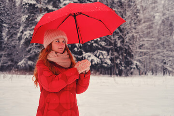 A woman with a red umbrella stands in a winter forest among trees in the snow. An adult happy woman with red hair takes shelter from the snow with an umbrella