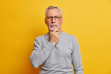 Senior thougthful man holds chin and looks pensively aside makes plannings wears spectacles and casual grey jumper isolated over vivid yellow background. Handsome grandfather considers something - Powered by Adobe