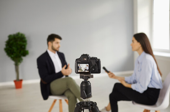 Digital Video Camera On Tripod Recording Interview With A Person In Television Studio. TV Show Host Talking To Celebrity. Journalist Or Reporter Asking Famous Businessman Questions. Blurred Background