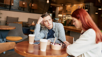 Two teenagers spending time together, sitting in a cafe on a daytime. Displeased guy looking at his girlfriend while she is ignoring him, using her smartphone