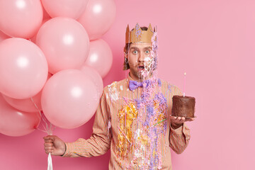 Indoor shot of stupefied man poses with balloons and cake wears paper crown on head being dirty...