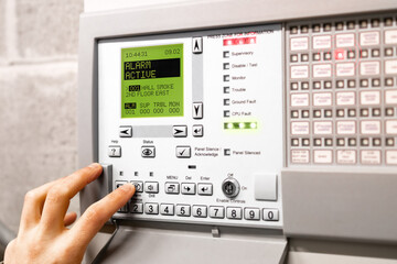 Fire alarm control panel is activated and in alert mode. Display message: Alarm active hall smoke. Red flickering lights and peeping. A hand is using the silence button. Selective focus. - obrazy, fototapety, plakaty