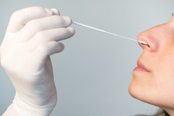 A nurse wearing latex gloves inserts a swab into a woman's nose to collect a possible positive...