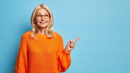 Thoughtful blonde forty years old European woman wears spectacles and knitted orange sweater points away on blank copy space shows place for your advertisement isolated over blue background. - Powered by Adobe
