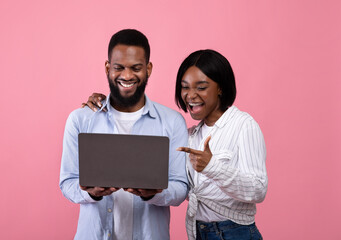 Black couple using laptop, surfing web, watching movie, pointing at screen, learning exciting news on pink background