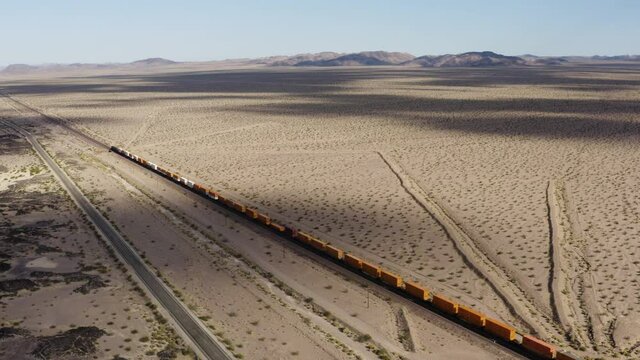 Freight train driving through the flat Californian dessert as clouds throw huge shadows on the landscape. Drone.