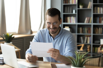 Smiling young businessman in eyeglasses looking at paper correspondence, reading pleasant good...