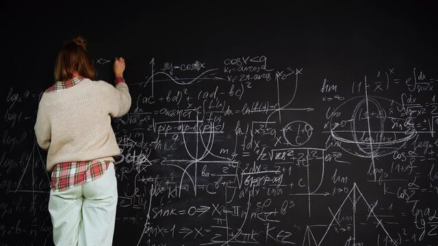 Woman solves scientific problem writing formulas on chalkboard focused on studies. Female smart student draws mathematical graphs and proves the theorem. Caucasian professor explains algebra material.