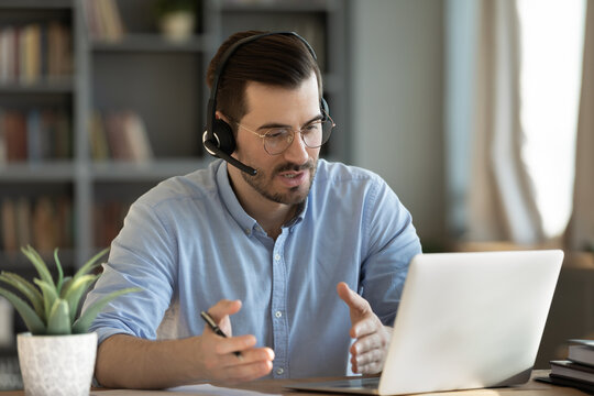 Concentrated skilled millennial caucasian businessman in glasses wearing headphones with mic, taking part in online web camera negotiations meeting using computer app, distant communication concept.