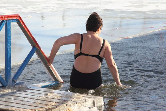 Winter Swimming Sport, A Mature Russian Woman In A Black Swimsuit Enters The Ice Hole Water On A Sunny Frosty Winter Day, Healthy Lifestyle