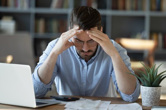 Frustrated young man in glasses sitting at table, feeling stressed calculating earnings or domestic expenses. Unhappy millennial guy suffering from lack of money, banking debt bankruptcy concept.