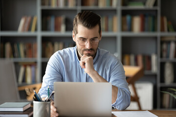 Thoughtful young 30s caucasian businessman in eyeglasses looking at computer screen, working...
