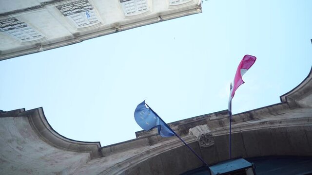 EU and French flag waving in the wind next to each other, looking up stone buildings and blue sky on a sunny day