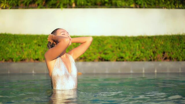 Slow motion shot of a attractive young woman standing half submerged in the pool touching her wet long hair with her eyes closed and then slowly opening them with delight and smile on her face.