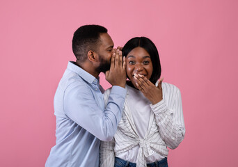 Naklejka premium Black guy sharing secret or whispering gossips into his girlfriend's ear on pink studio background