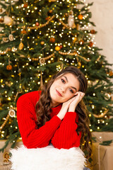 young beautiful girl in a red sweater and blue jeans sits near a Christmas tree in a cozy room decorated with Christmas decoration