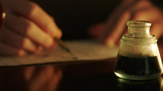 Close up of the hands of a man using an 18th writing implement that is dipped in ink from an inkwell and that is called a dip pen.