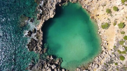 Stunning aerial descending view of a natural rock pool with crystal clear green water surrounded by rough nature and rolling waves of the open ocean