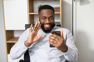 Cheerful young african-American guy using smartphone for video call, virtual meeting, conversation online indoor. A smiling multiracial man glad to talk via video