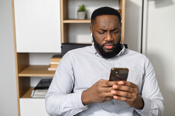 Obraz na płótnie Canvas Pensive puzzled African-American man in smart casual shirt holding smartphone and expressing misunderstanding, texting message, answering email indoor