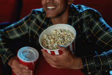 Cropped portrait of cheerful young guy smiling, holding a drink and popcorn basket while watching movie alone in empty theater auditorium