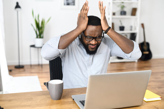 Frustrated young African-American businessman in smart casual holding head in hands, sitting alone at the modern office desk with an open laptop, stressing out from occurred computer issues