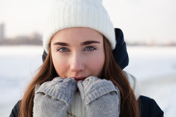 Winter portrait photo of young woman. Outdoor photo of long-haired happy lady in knitted hat having fun in snowy morning on blur nature background. Woman looking at the camera. Woman in gloves
