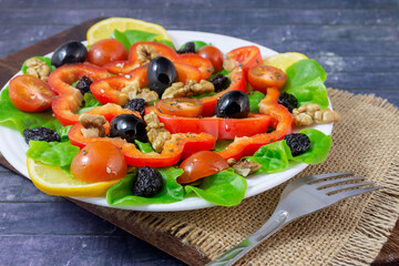 On a white plate, fresh vegetable salad with cherry tomatoes, olives, red peppers, walnuts, dried raisins, lettuce leaves.