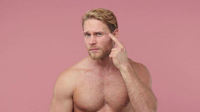 Bearded naked young man 20s perfect skin isolated on pink color background studio. Skin care healthcare cosmetic procedures concept. Playing with his face touching pointing finger looking for wrinkles