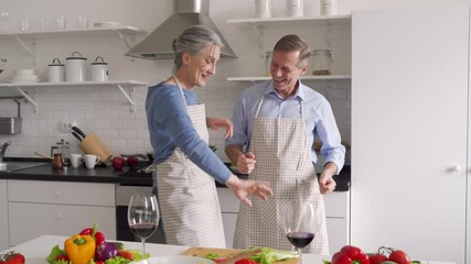 Happy affectionate older middle aged couple laughing, dancing, preparing healthy meal in modern kitchen apartment. Senior husband and wife enjoying dance having fun celebrating Valentines day at home. - Powered by Adobe